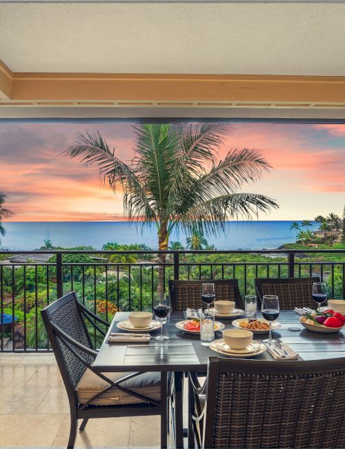 Balcony dining table with wine glasses and food, ocean and palm trees at sunset, plus a built-in grill beside the patio.