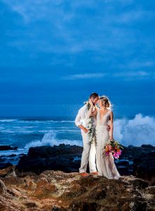 A wedding couple stands on rocky shore by the ocean, kissing as waves splash behind them.