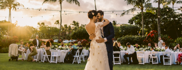 A bride and groom dance at an outdoor wedding on a tiled dance floor, with guests seated behind them under string lights and palm trees.