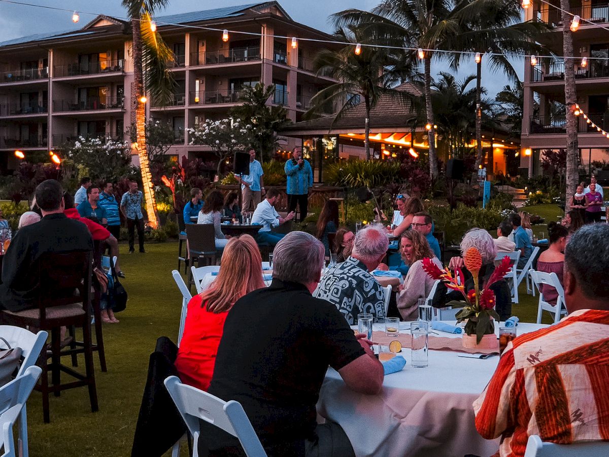 An outdoor party at a tropical resort courtyard with string lights and guests dining at tables, while staff speak near the stage.