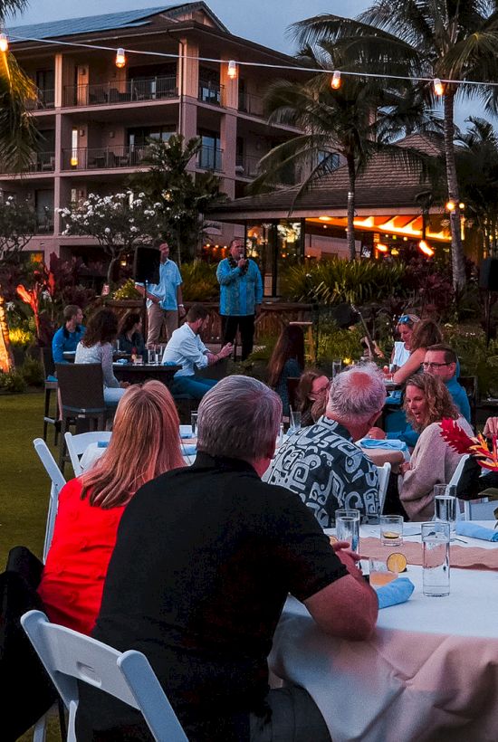 An outdoor party at a tropical resort courtyard with string lights and guests dining at tables, while staff speak near the stage.