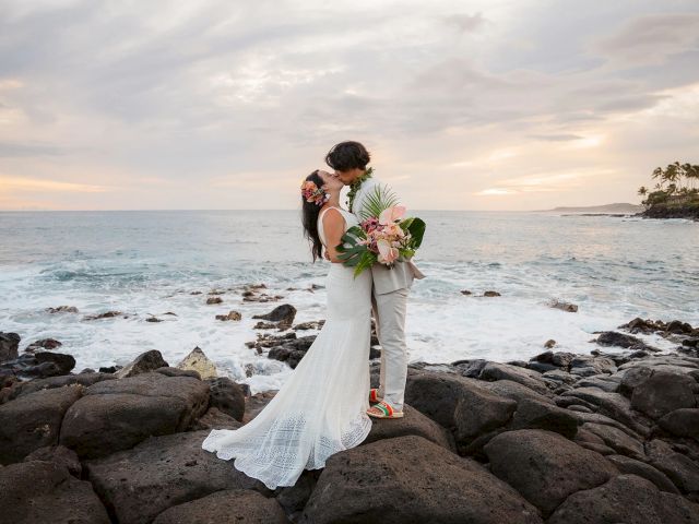 A couple in wedding attire kisses on a rocky beach at sunset, holding a bouquet, with the ocean behind them.