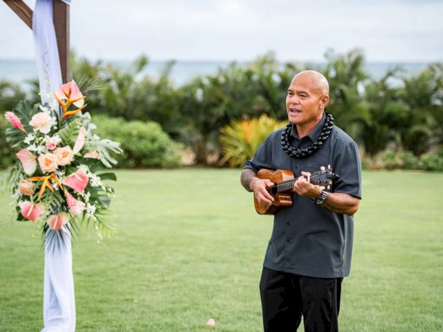 An outdoor ceremony: a bald man in a lei plays a ukulele while a colorful wedding bouquet stands beside him.