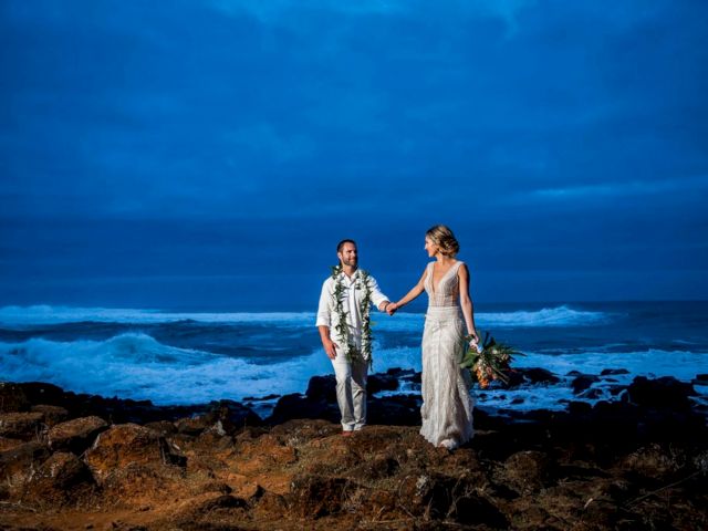 A couple in wedding outfits stands on rocky shore, holding hands as waves crash under a dramatic blue sky.