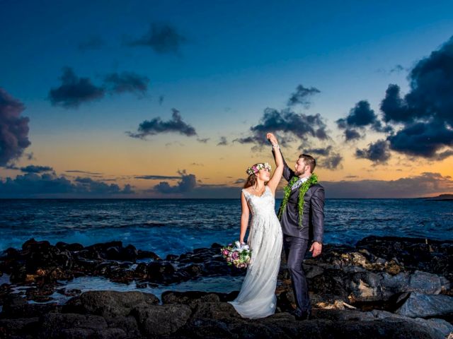 Bride and groom pose on a rocky beach at sunset, with the groom raising the bride&rsquo;s hand.