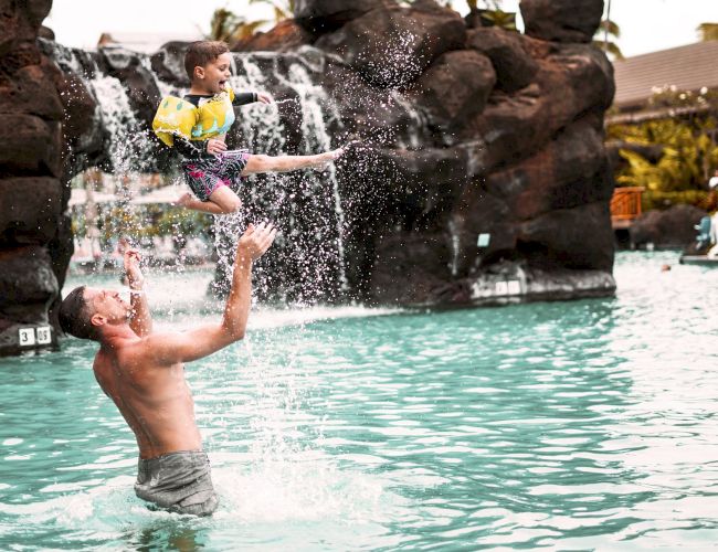 A man lifts a child midair above a pool as water splashes.
